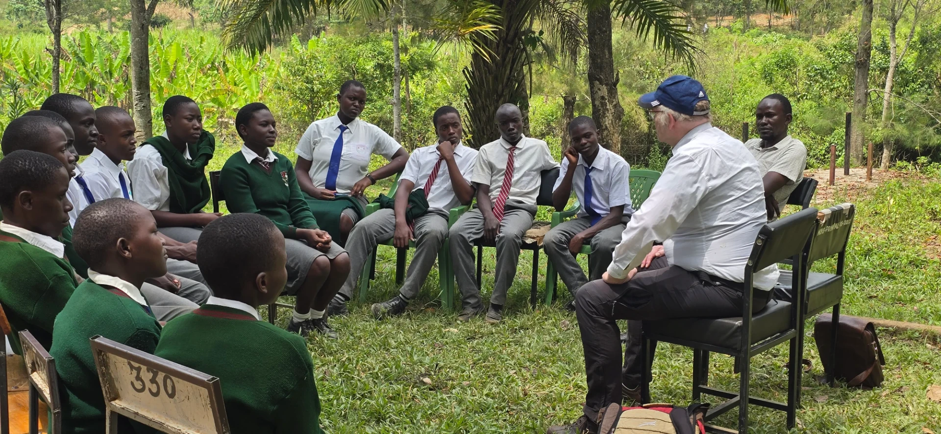 Sponsored students in green and white uniforms sit in a circle outdoors discussing with a visitor